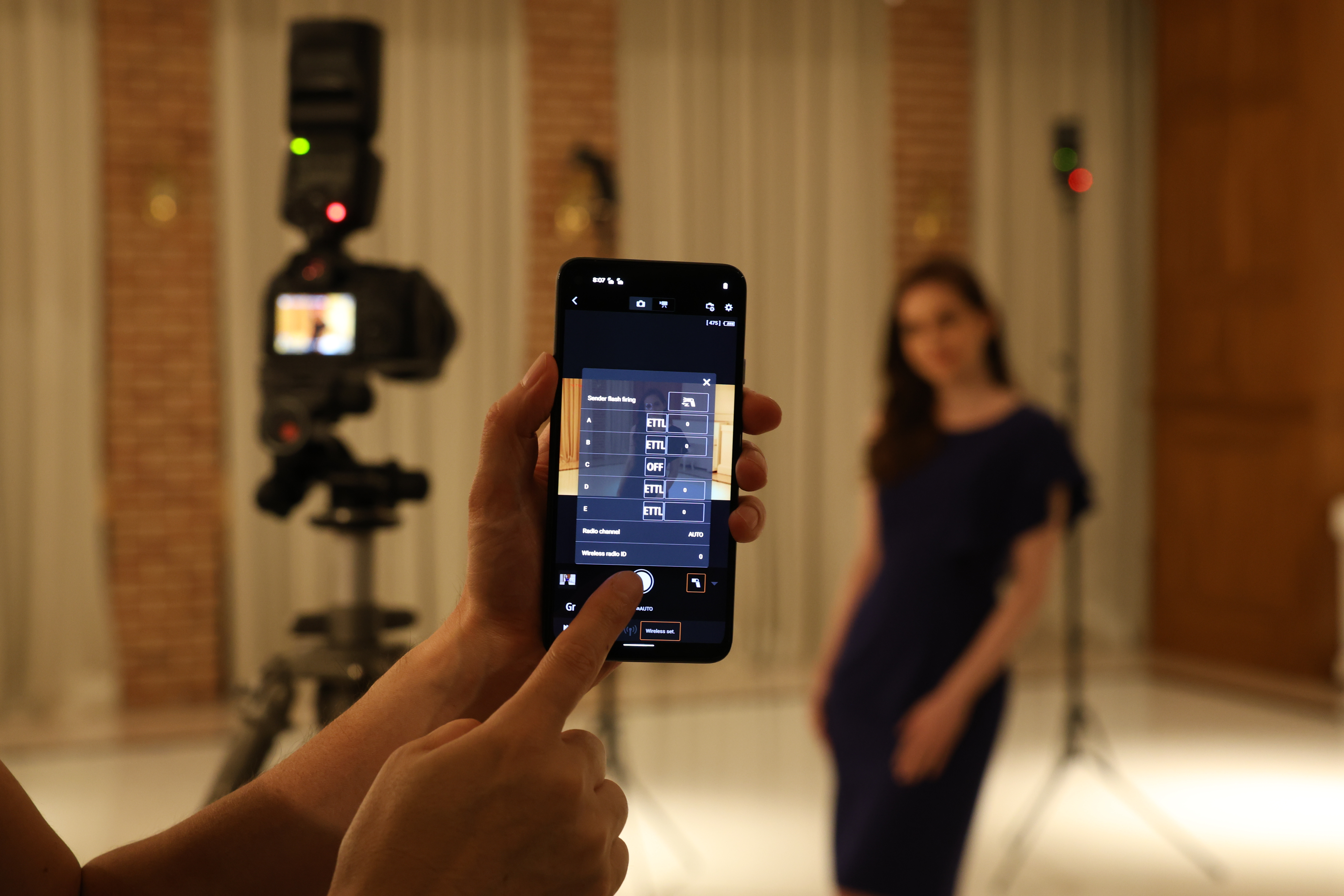 Eine Person hält ein Telefon und richtet eine Kamera für ein Model in einem blauen Kleid ein. Ein Blitz steht auf einem Stativ.