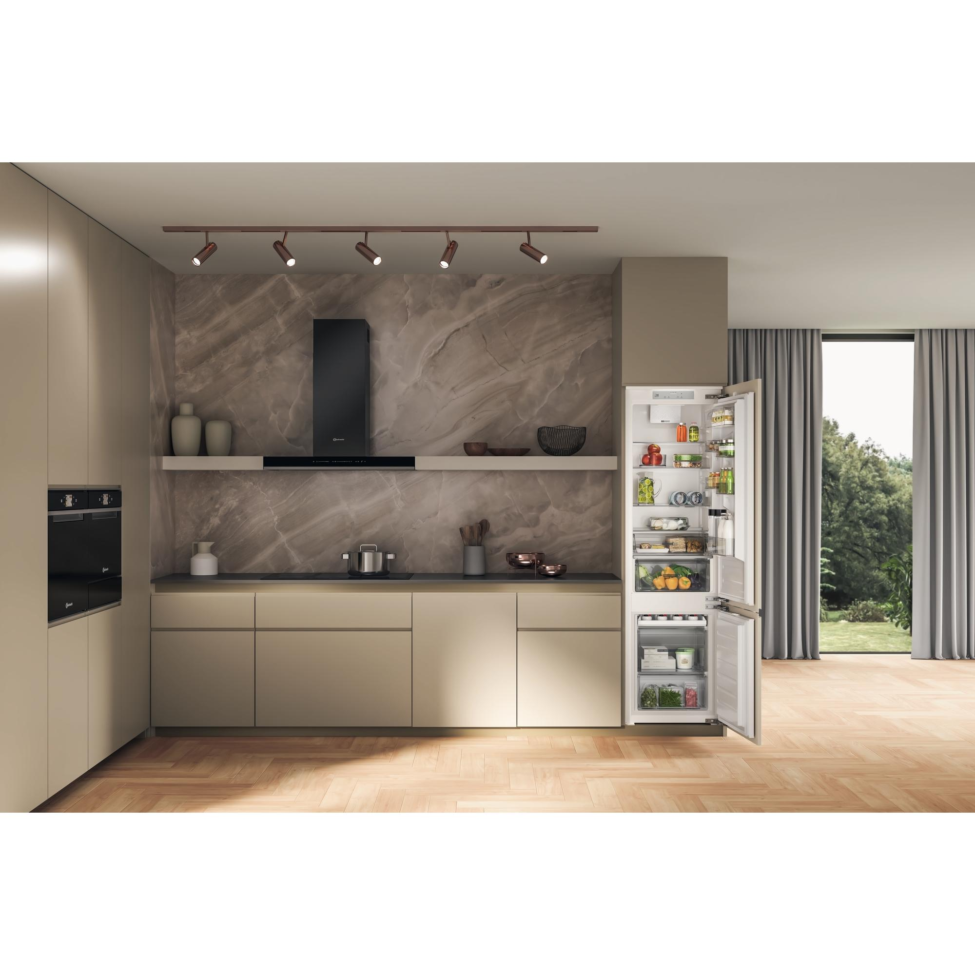 Küche mit geöffnetem Kühlschrank, gefüllt mit Lebensmitteln. Deckenleuchten, Holzschränke und Fenster.