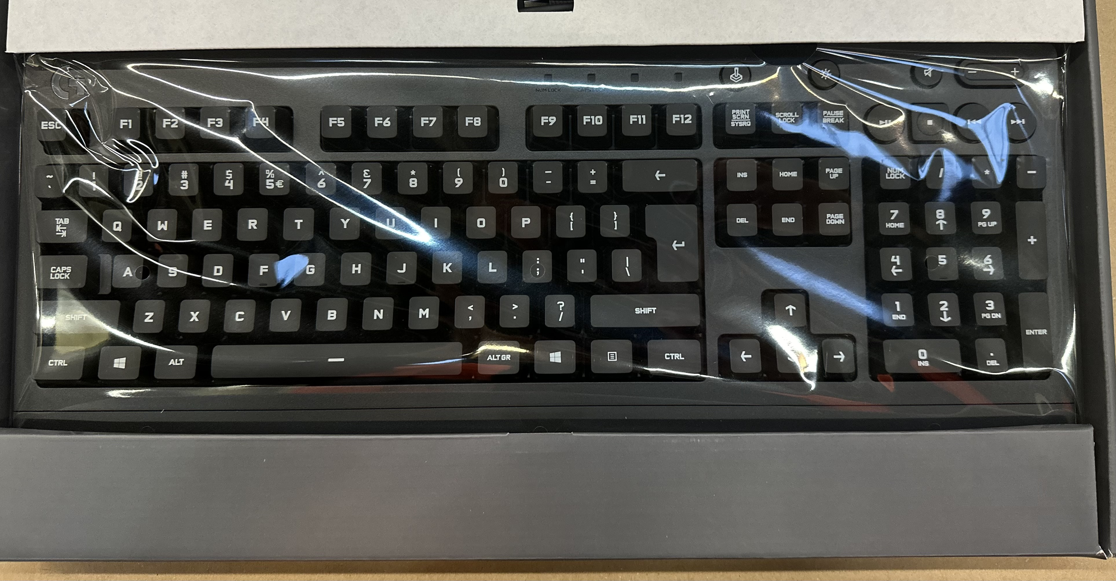 Czarna klawiatura w przezroczystym plastikowym opakowaniu, w szarym pudełku, widoczne klawisze.