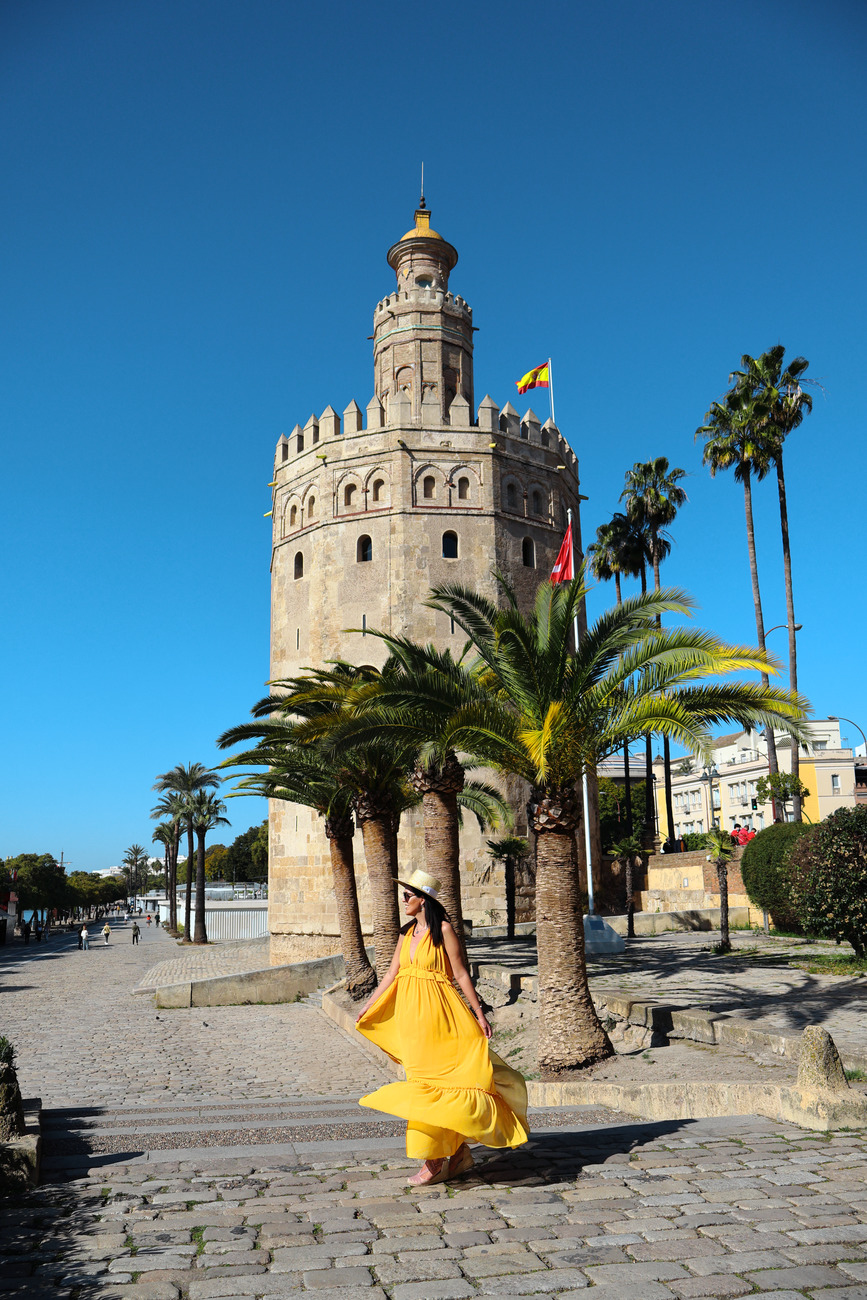 Frau im gelben Kleid steht bei Turm, Palmen, sonniger Tag.
