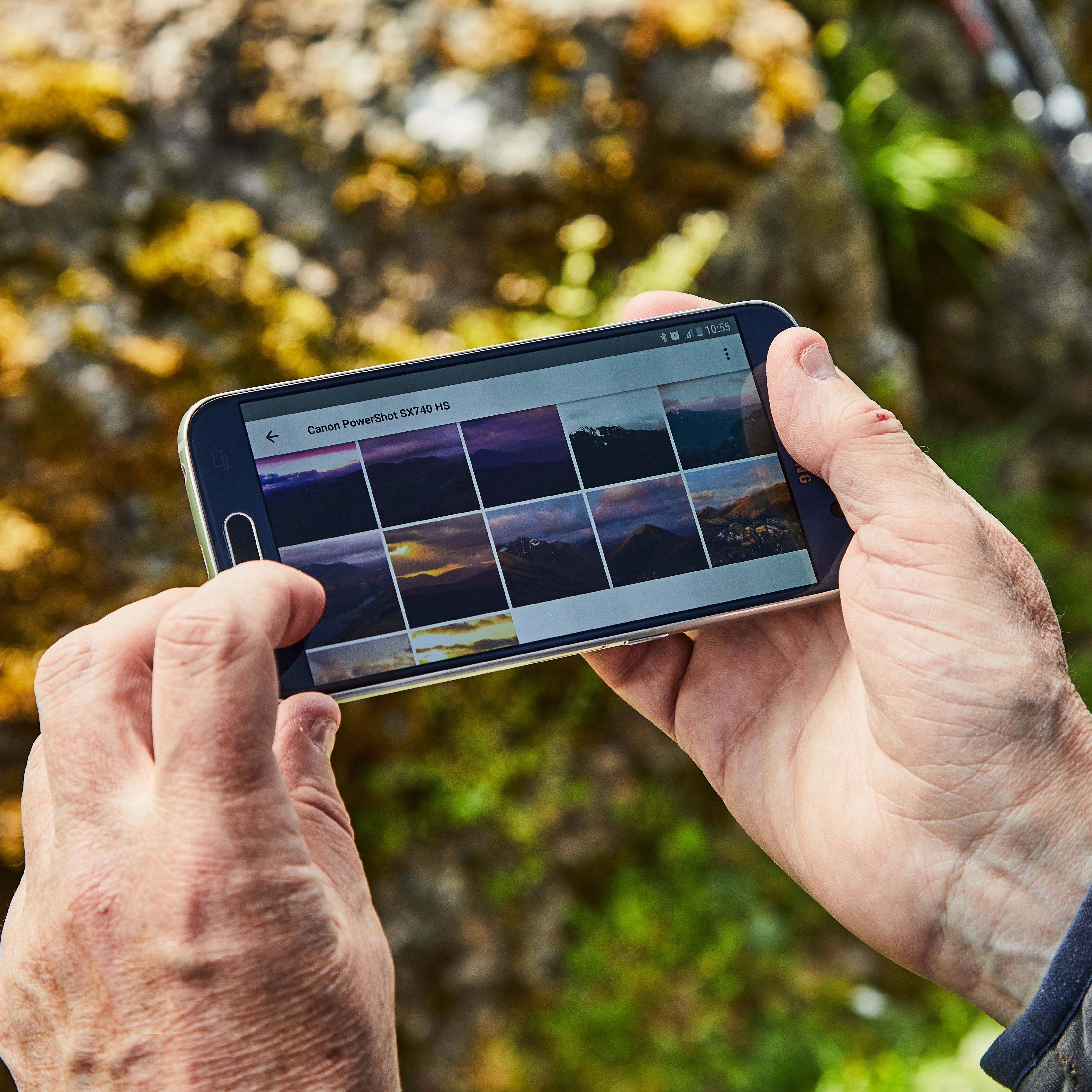 Hände halten ein Telefon mit Fotos von Bergen. Der Hintergrund ist unscharfer Felsen und Blätter.