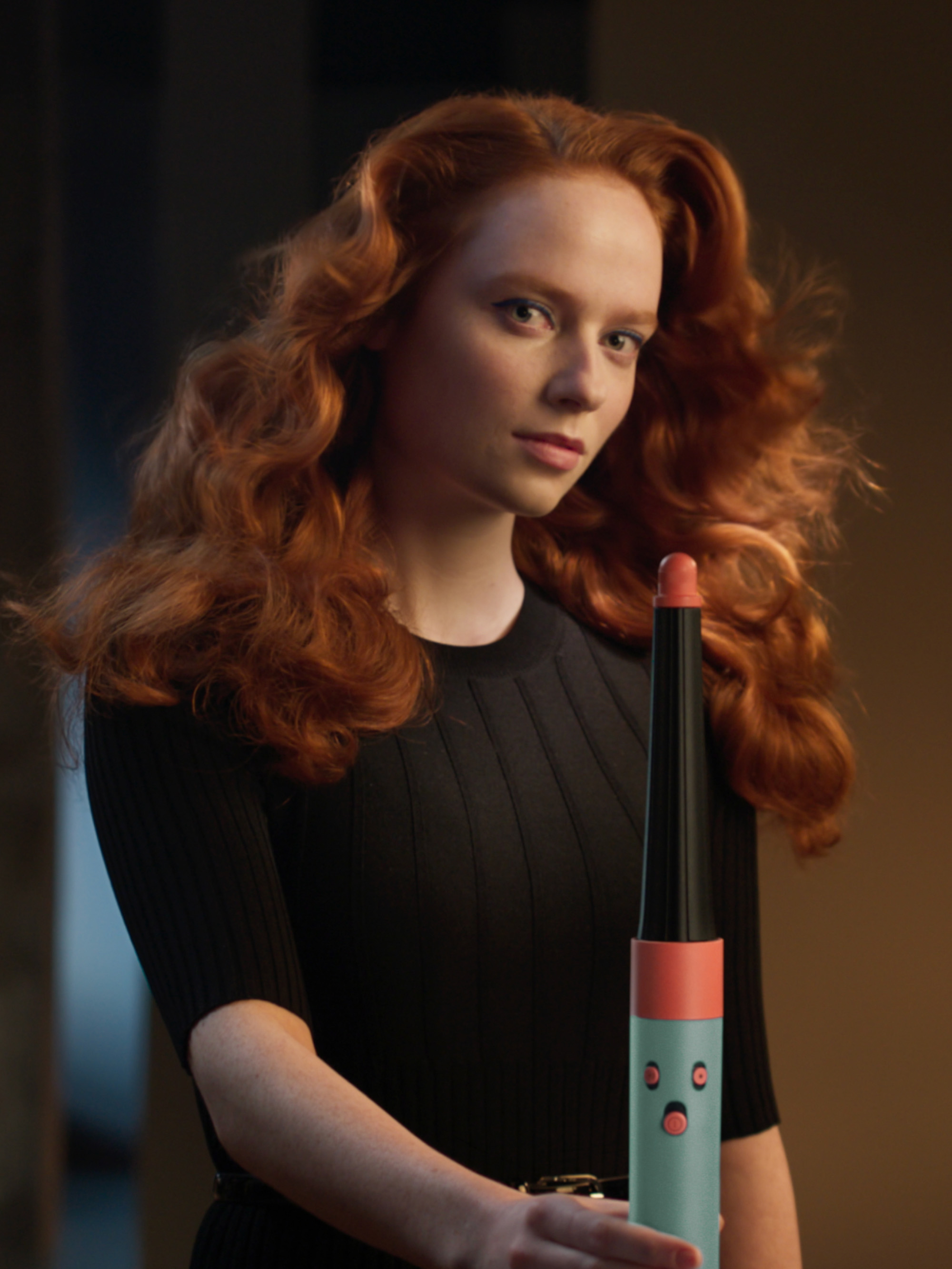 Una mujer con cabello rojo sosteniendo un rizador de pelo turquesa, con un vestido negro y un fondo oscuro.
