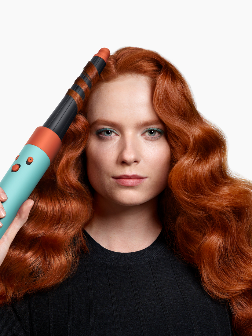 Una mujer con cabello rojo está usando un rizador. La plancha es turquesa y naranja.