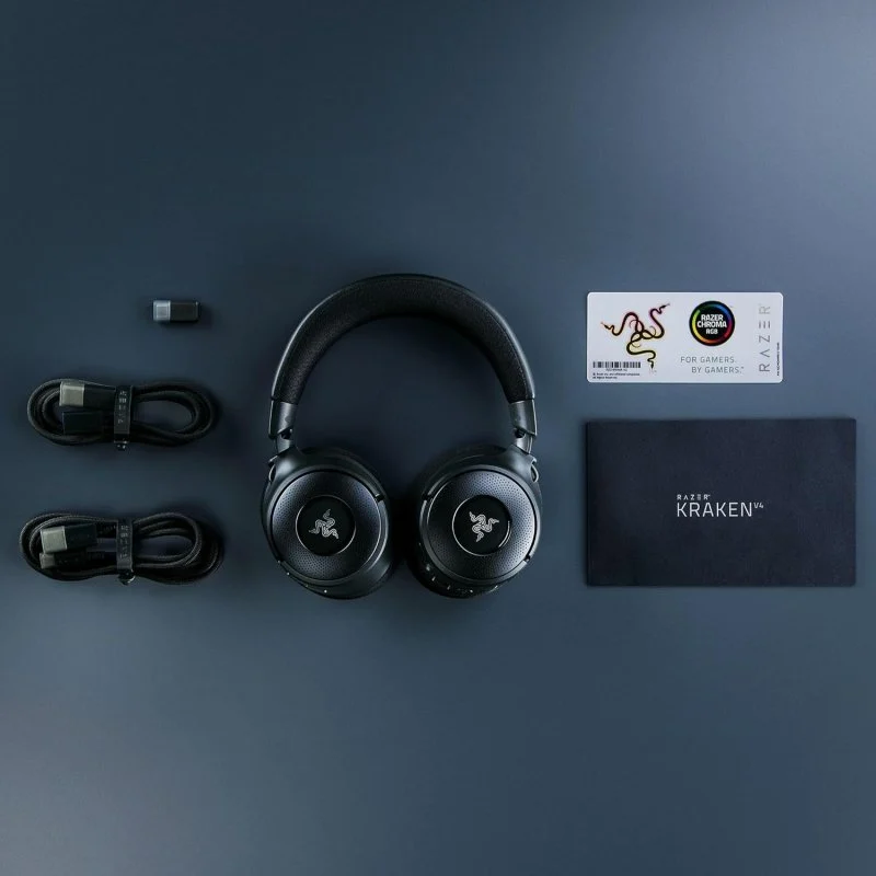 Auriculares Razer Kraken V3 negros, cables, dongle, tarjeta y caja sobre superficie azul oscuro. Logo Razer en auriculares.