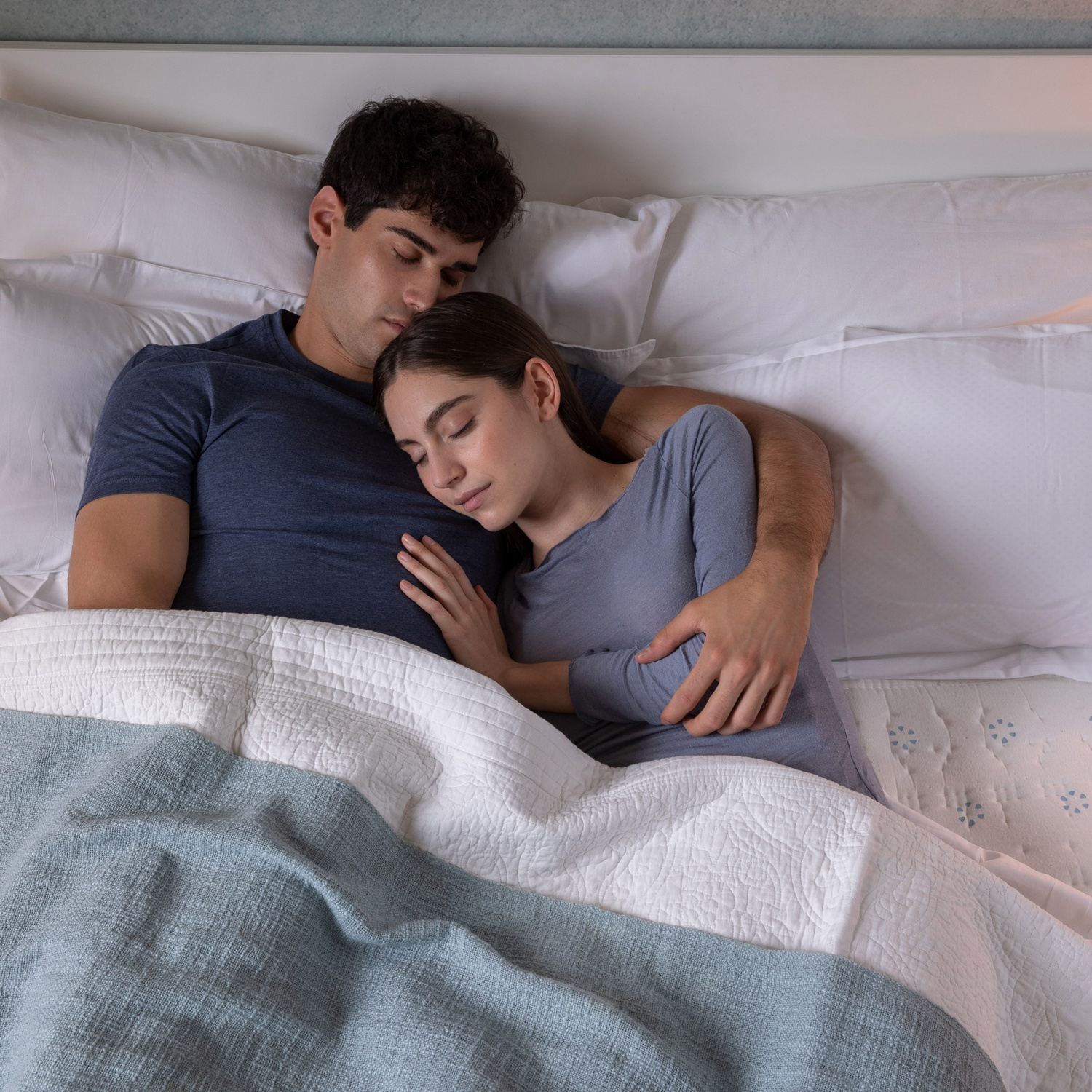 Una coppia che dorme in un letto, coperta da un piumino bianco e una coperta blu.