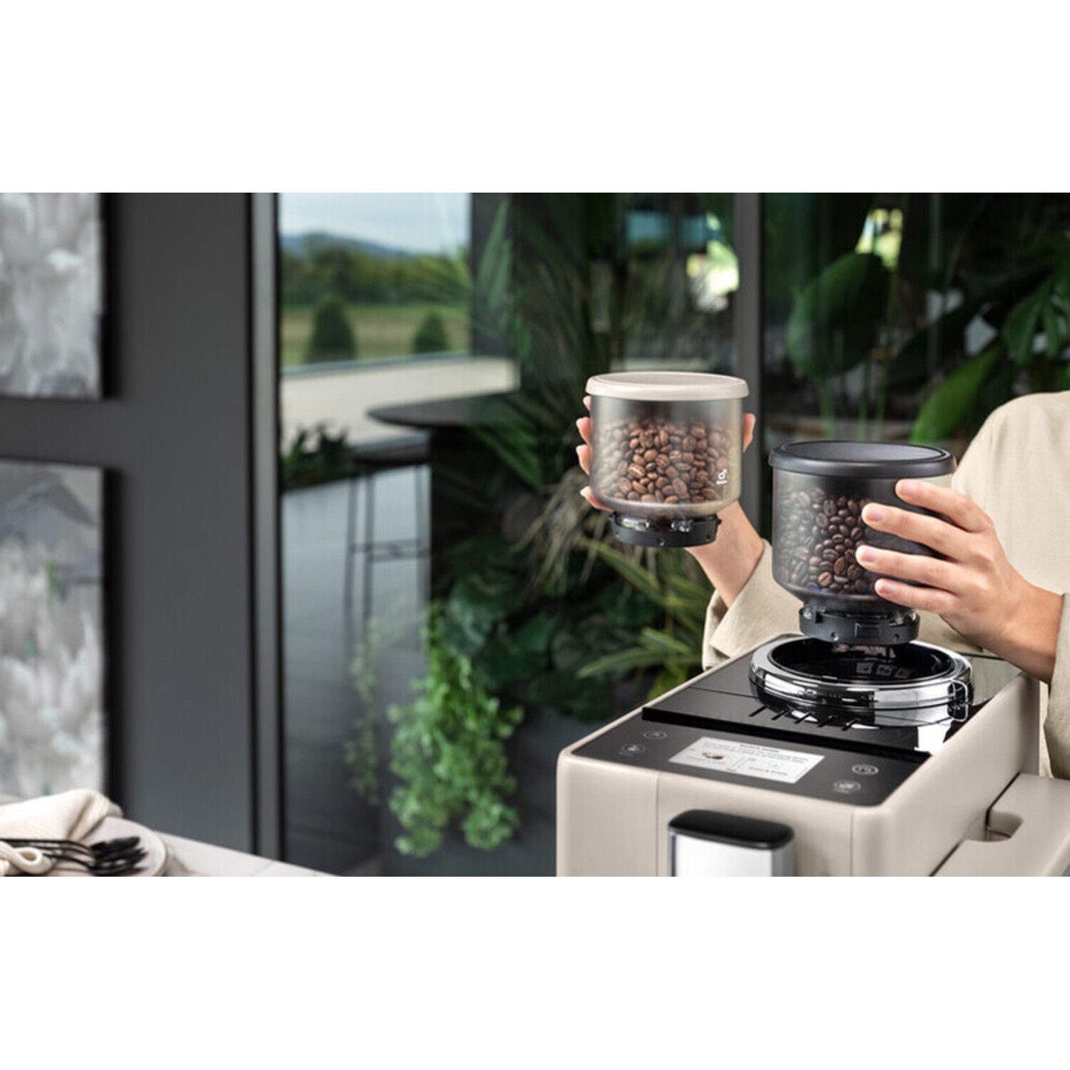 A person holding two containers of coffee beans above a coffee machine. The containers are clear.