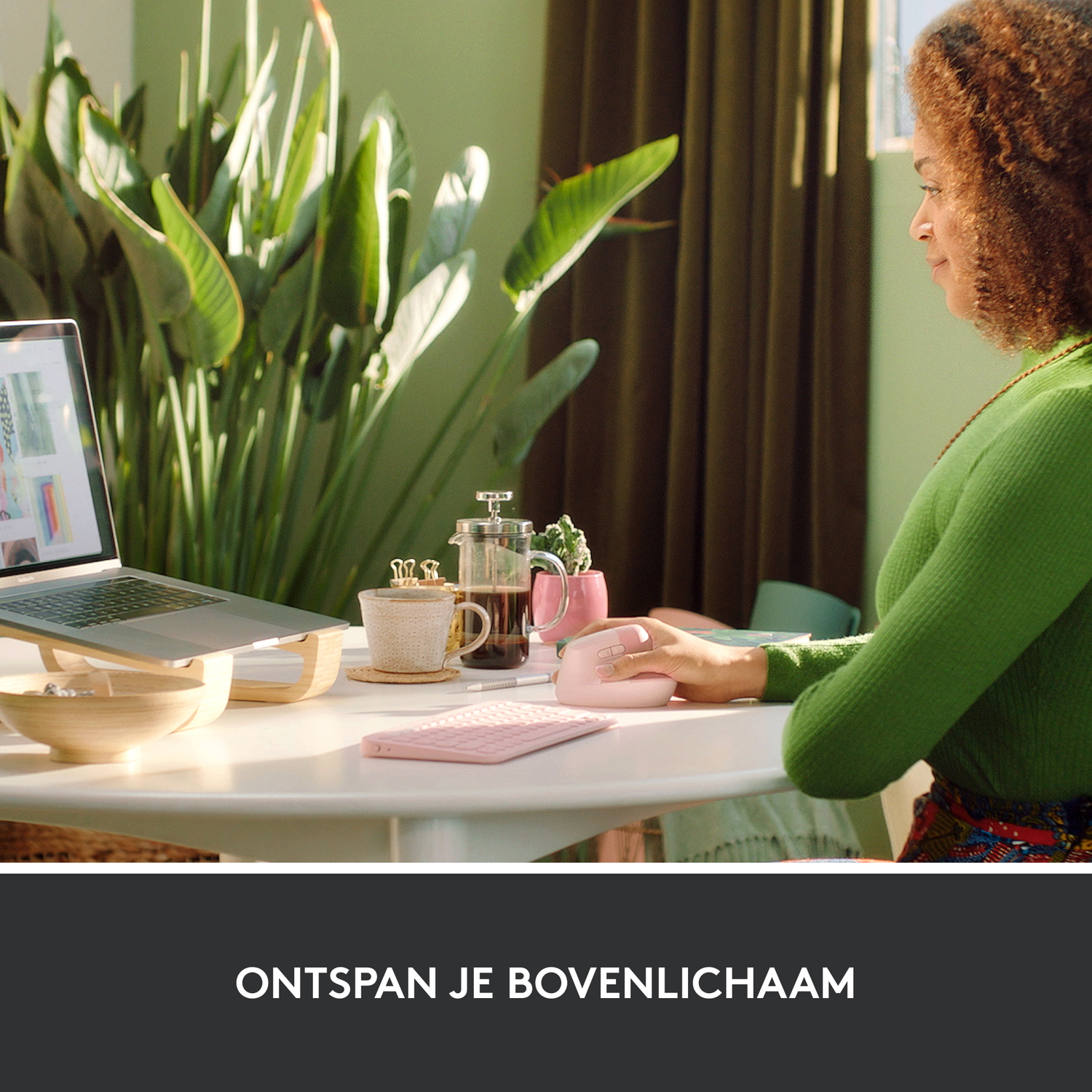 Woman in a green shirt is sitting at a table with a laptop, pink mouse and keyboard.