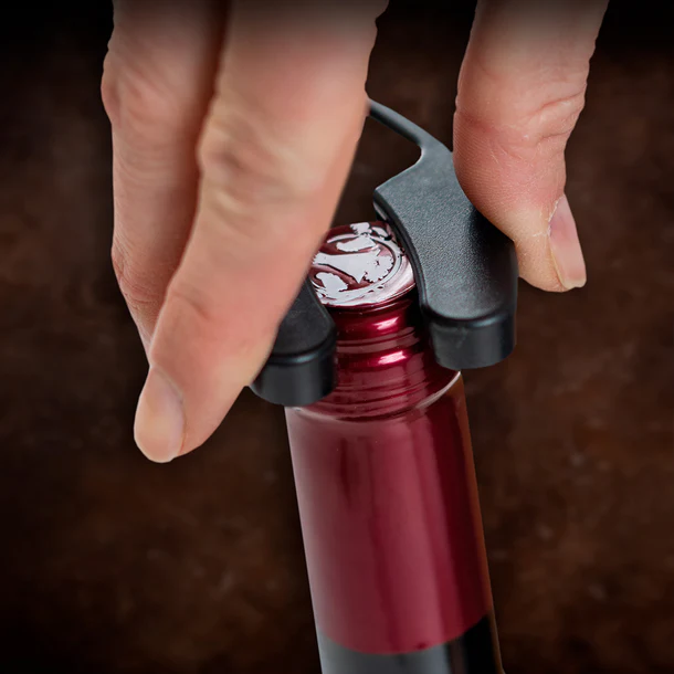 Una mano usa un abridor negro en una botella de vino tinto. El fondo es un marrón borroso.