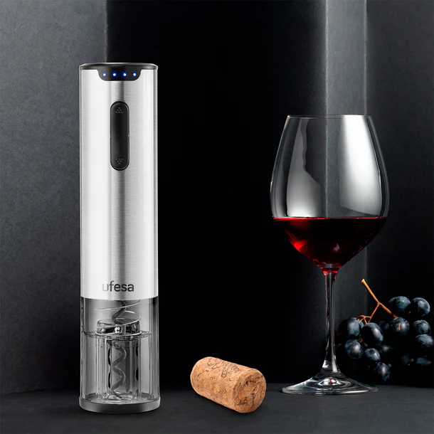 Abridor de vino eléctrico plateado, copa de vino tinto, corcho y uvas sobre fondo negro.