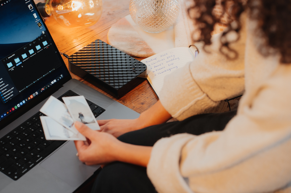 Osoba trzymająca zdjęcia obok laptopa z dyskiem twardym i notatnikiem, na drewnianym stole.