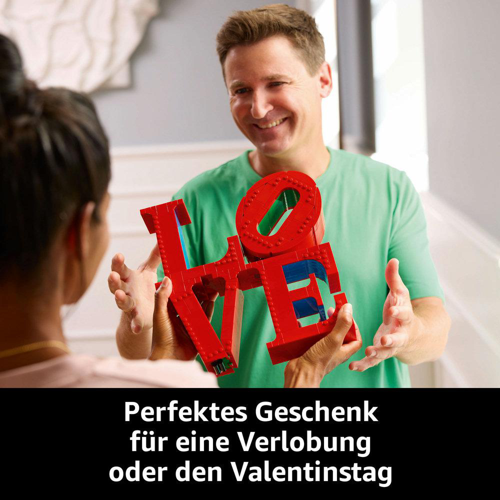 Ein Mann hält ein rotes Lego LOVE-Schild. Eine andere Person steht ihm gegenüber. Weiße Tür im Hintergrund.