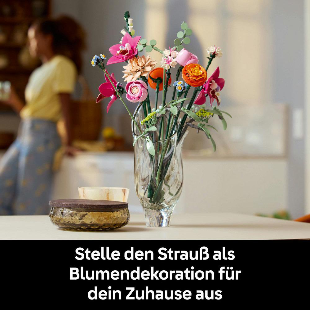 Eine Vase mit bunten Lego-Blumen steht auf einem Tisch. Im Hintergrund ist eine Frau. Der Text ist auf Deutsch.