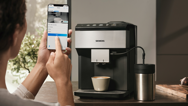 Kişi, kahve makinesini başlatmak için telefon kullanıyor. Kahve fincanı, paslanmaz çelik kap ve Siemens makinesi görünüyor.