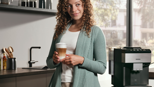 Mutfakta latte tutan kadın. Sağda Siemens kahve makinesi. Arka planda mutfak.