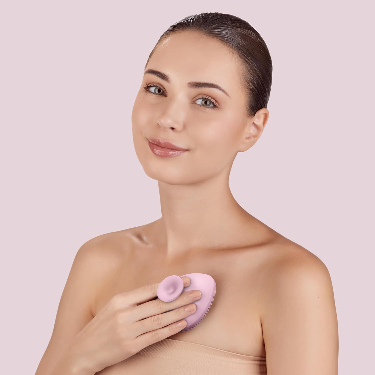 Mujer usando un masajeador facial rosa, sosteniéndolo cerca de su pecho, sobre un fondo rosa.