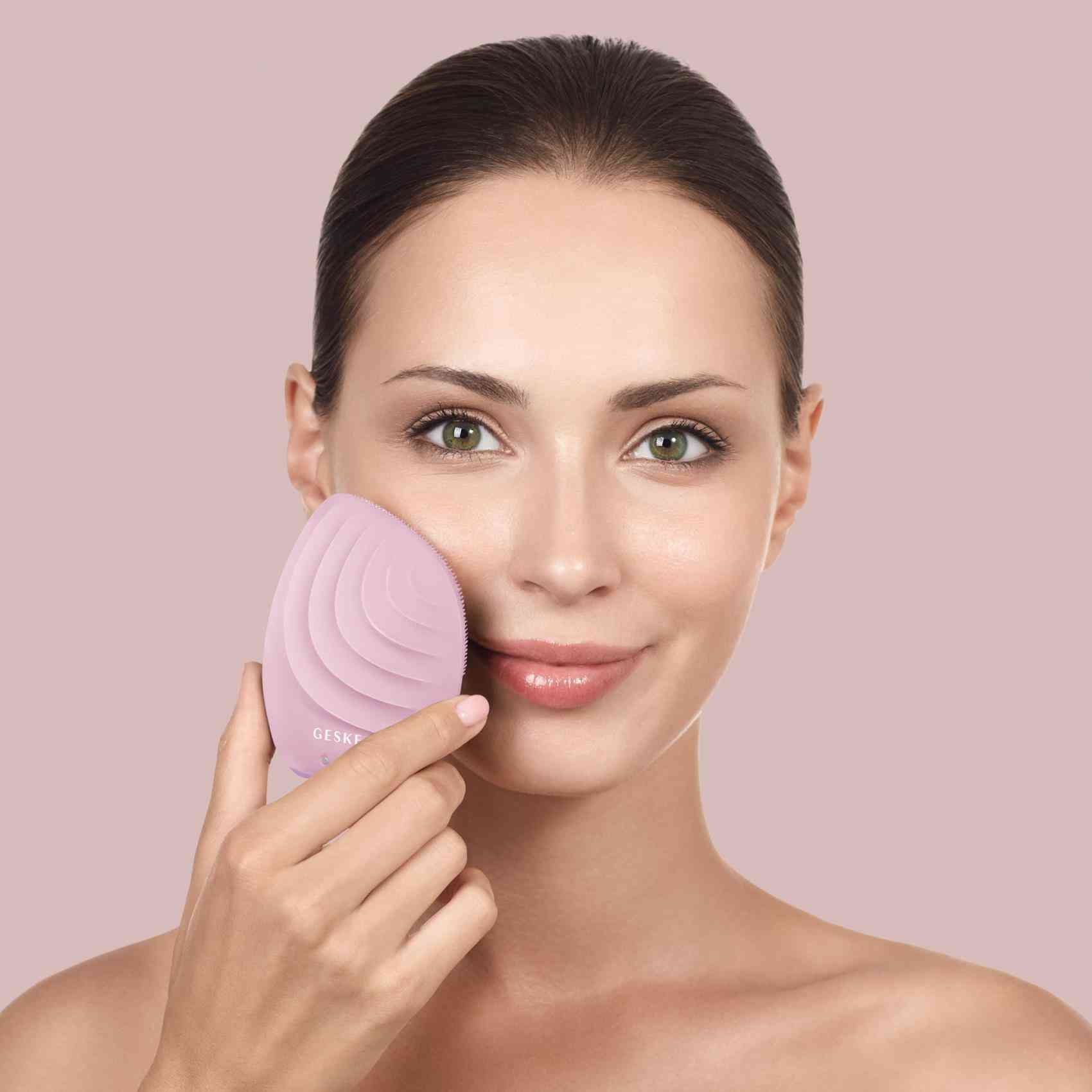 Una mujer usando un dispositivo de limpieza facial rosa en su cara, mirando a la cámara.