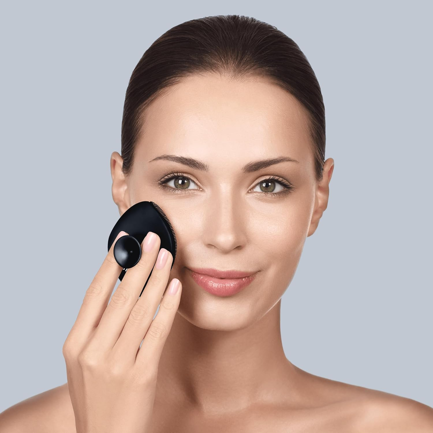Mujer usando un cepillo facial negro, mirando a la cámara, sobre un fondo gris.