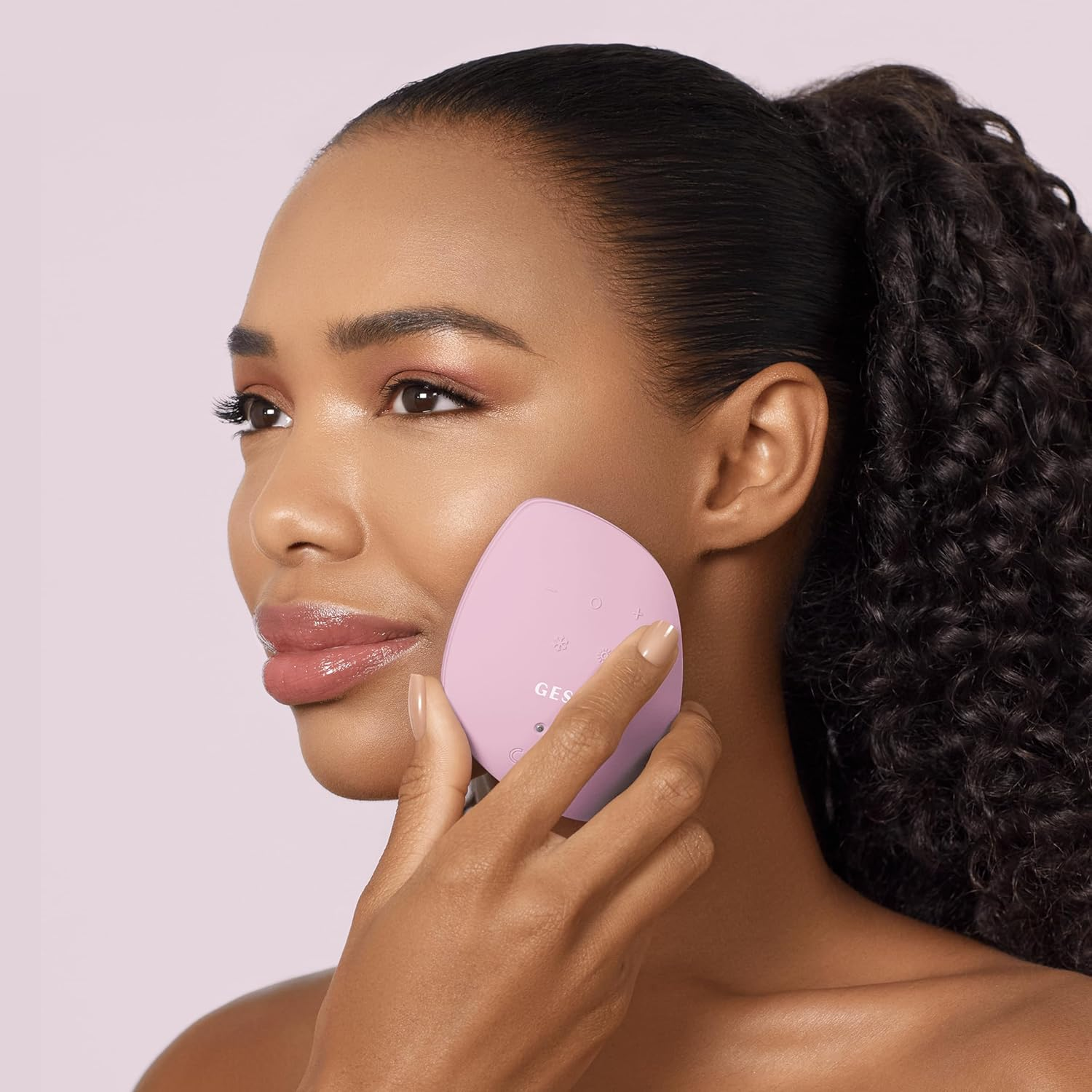 Mujer usando un dispositivo facial rosa, sosteniéndolo en su mejilla, sobre fondo rosa.