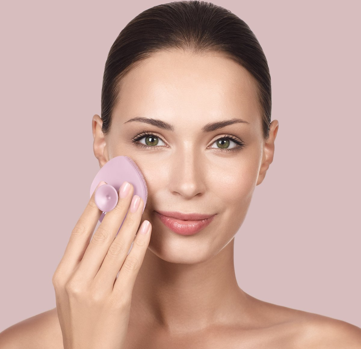 Una mujer aplica un cepillo limpiador facial rosa en su mejilla.