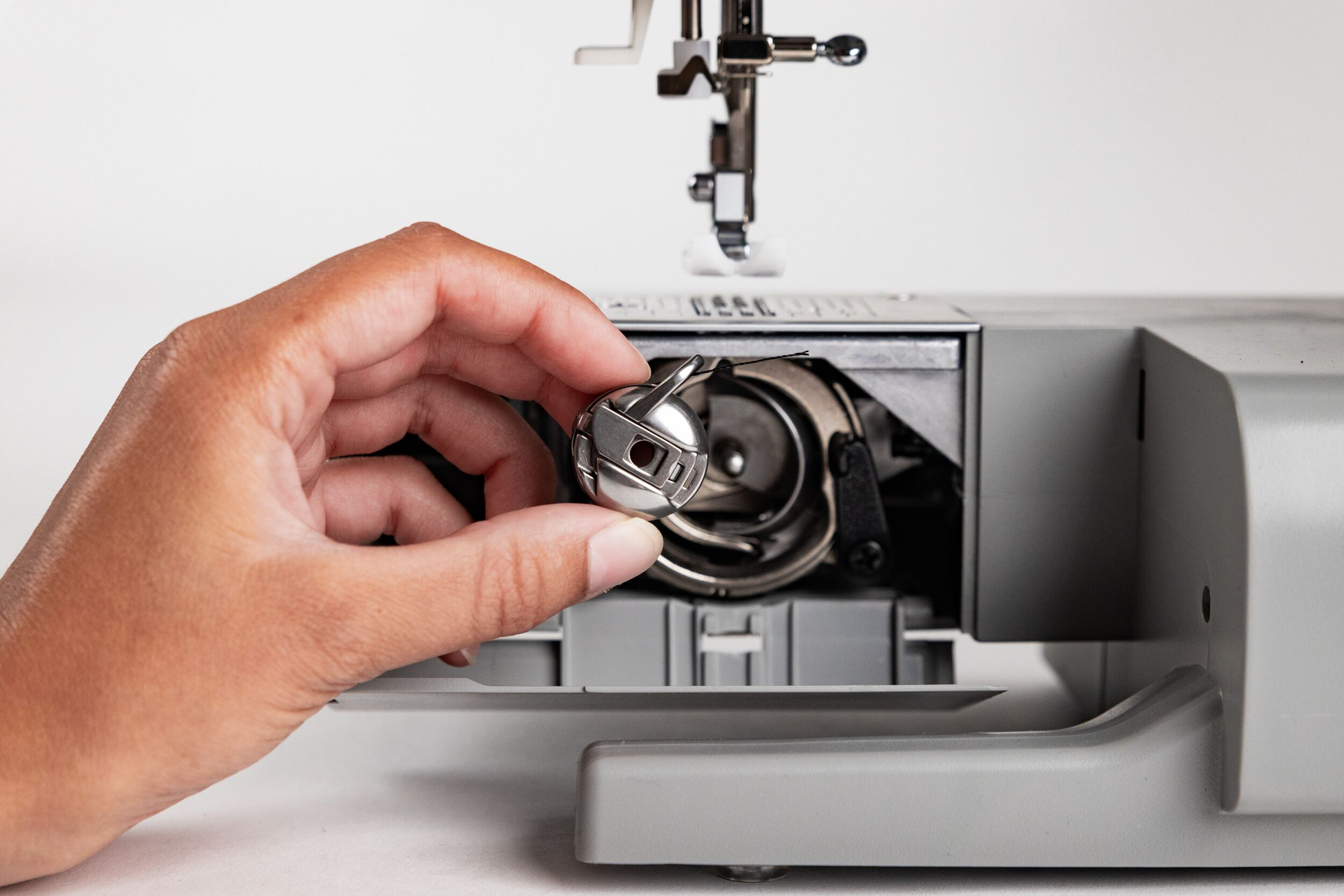 Hand hält eine Spule in der Nähe einer Nähmaschine. Die Maschine ist grau, mit geöffnetem Spulenkapsel.