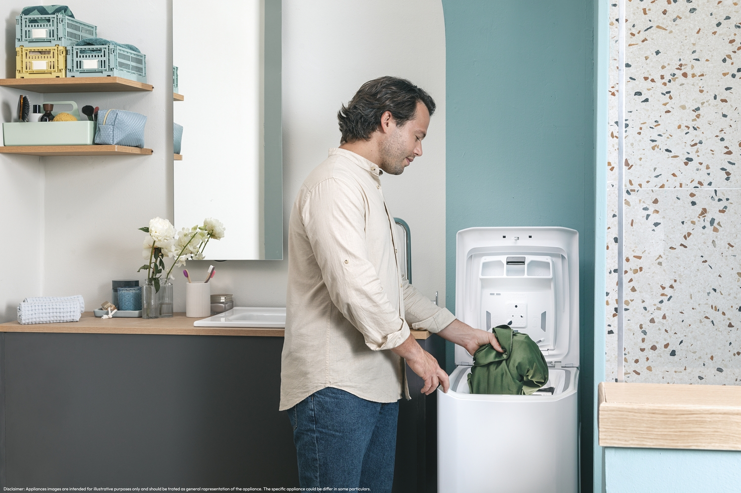 Un hombre poniendo ropa en una lavadora de carga superior blanca en un baño. Hay un lavabo, espejo y decoración a la vista.