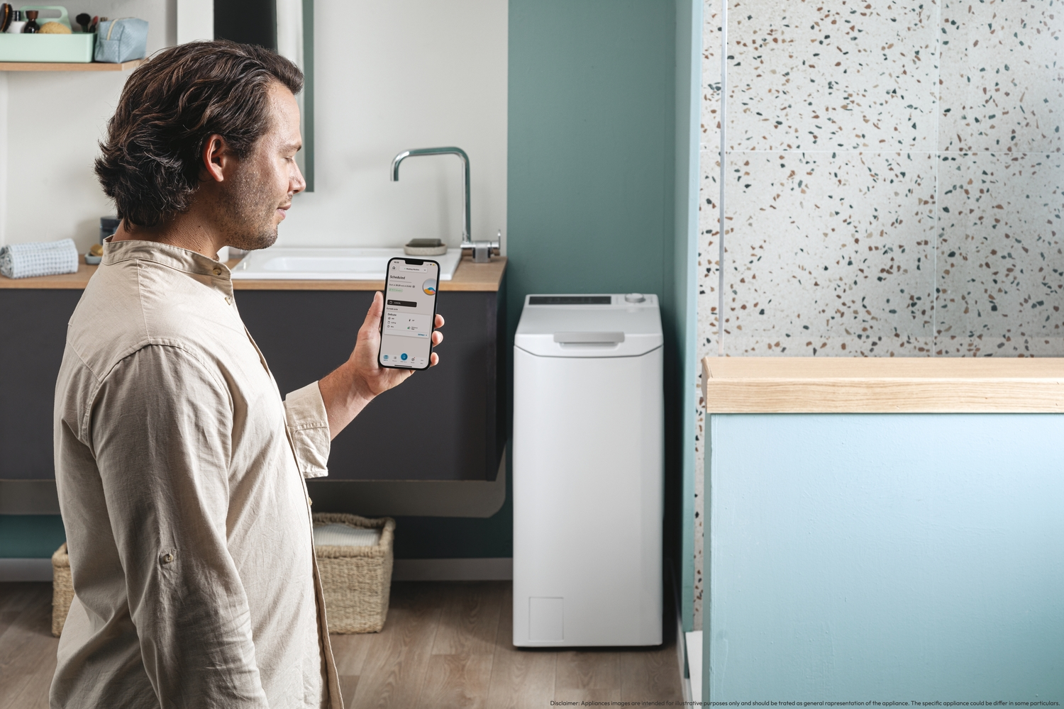 Un hombre sosteniendo un teléfono frente a una lavadora. Está en un baño, junto a un lavabo. Luz del día.