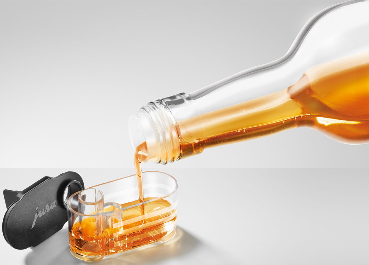 Eine Flasche gießt bernsteinfarbene Flüssigkeit in einen durchsichtigen Behälter. Links ein schwarzes Objekt mit "jura".