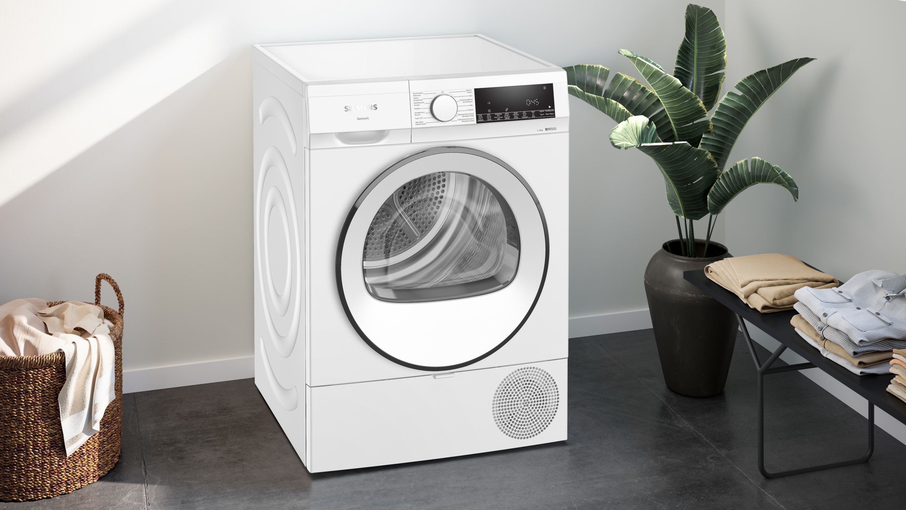 White dryer with a display, a basket of clothes, and a plant in the background.