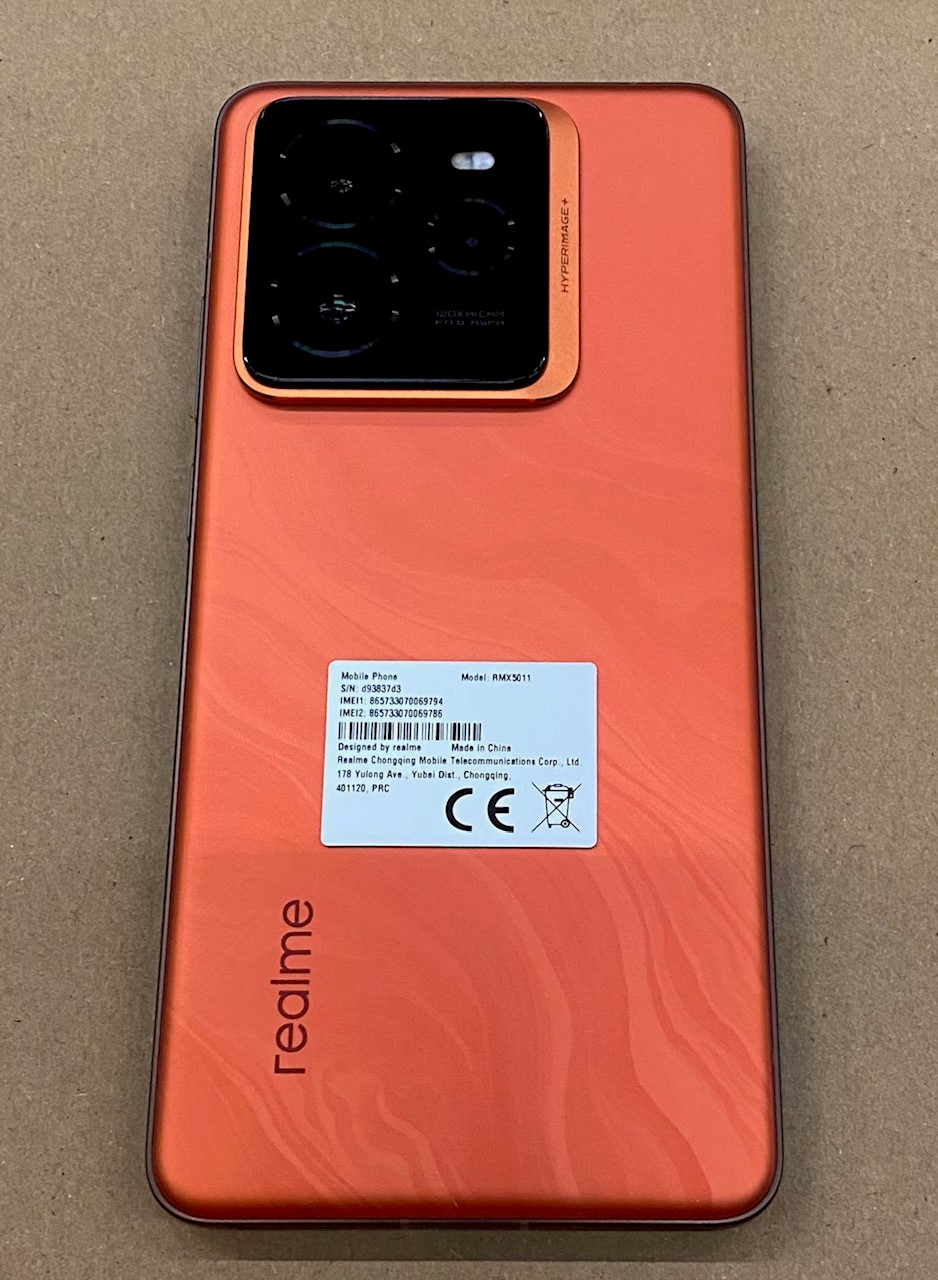 Pomarańczowy telefon Realme na brązowej powierzchni, z potrójnym aparatem z tyłu.