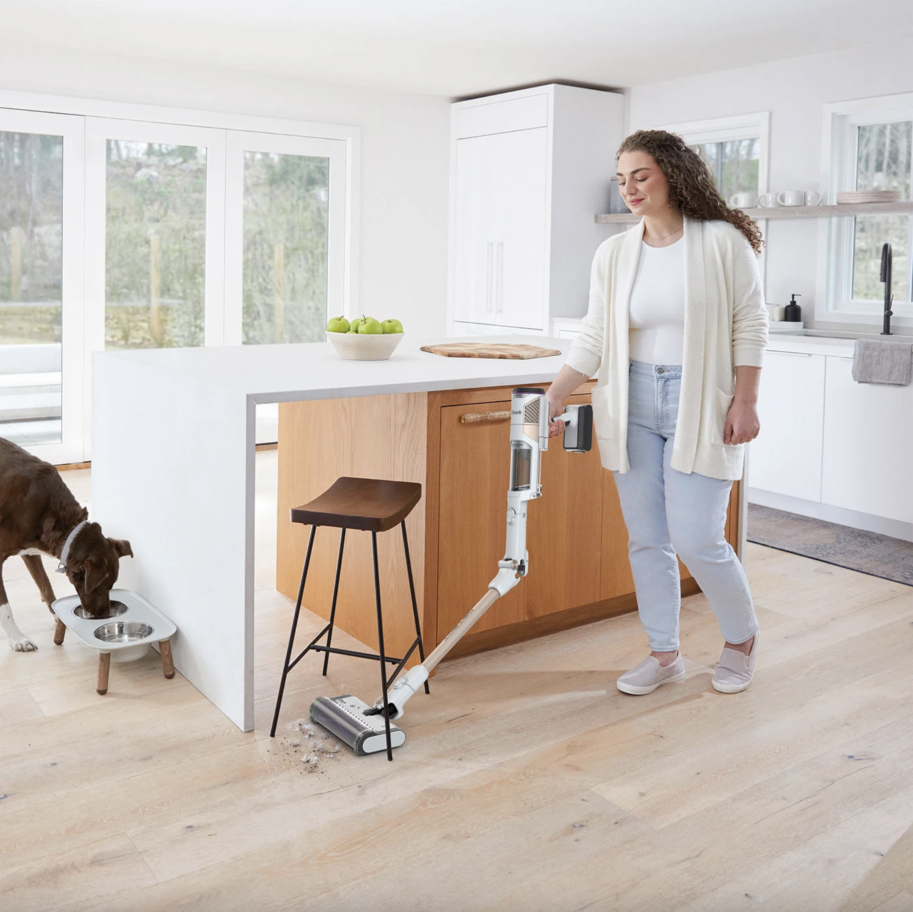 Femme passant l'aspirateur dans une cuisine avec un chien qui regarde. Un bol pour chien et un tabouret sont présents.
