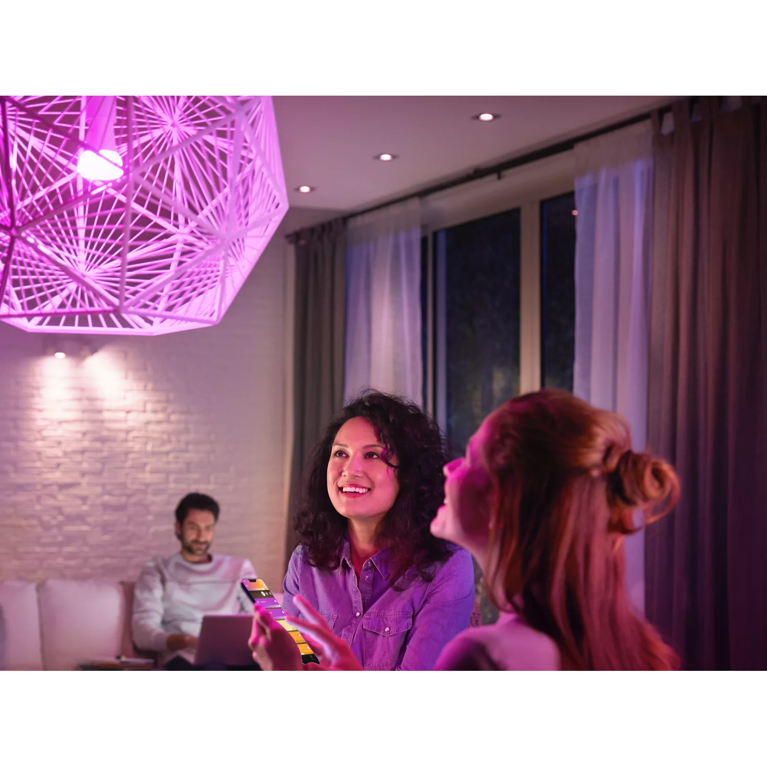 Two women and a man in a living room with pink lighting using a phone.
