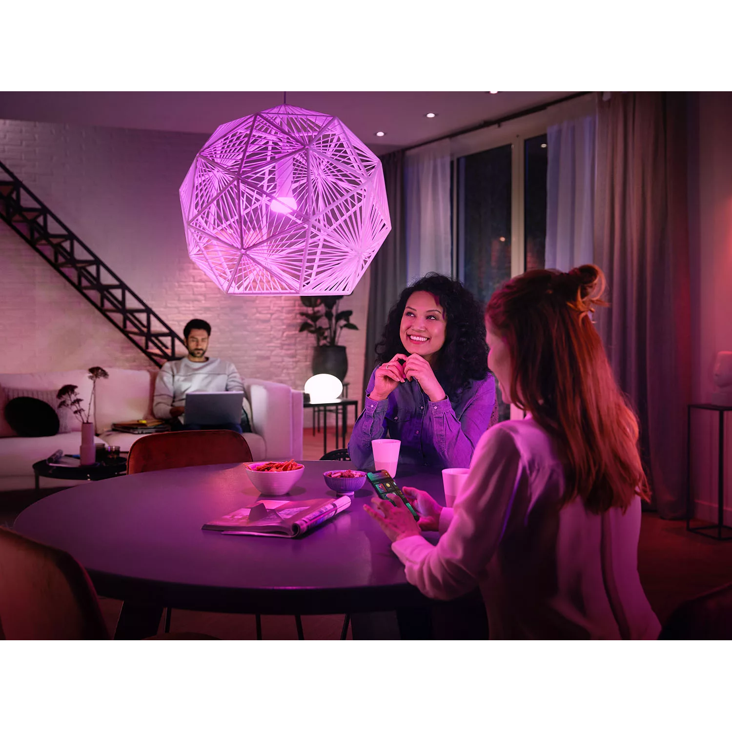 A brightly lit room. Two women are talking at a table. A man sits on a sofa. A large, geometric light fixture.