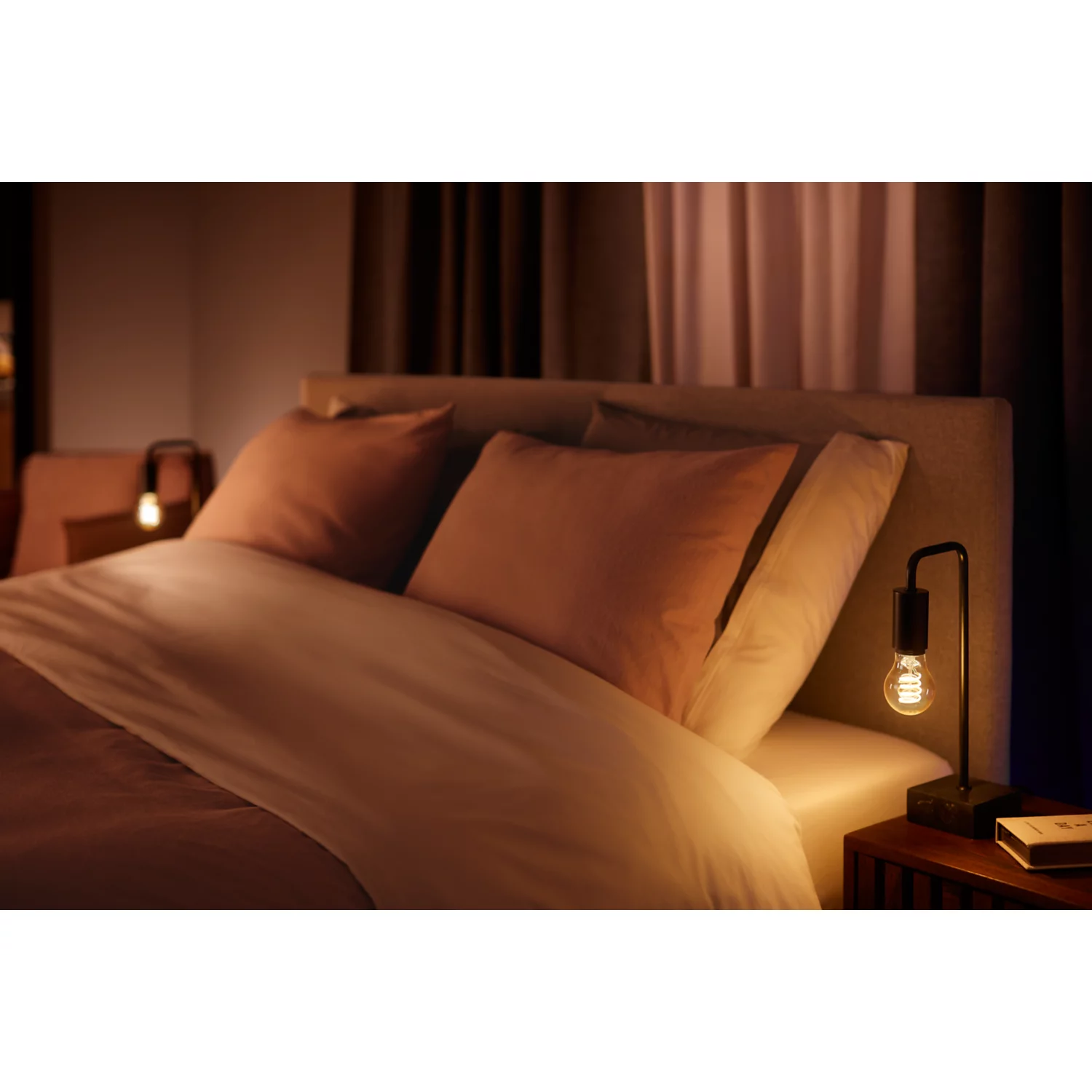 Bedroom with a bed, a lamp on a nightstand, and warm lighting. A book is also present.