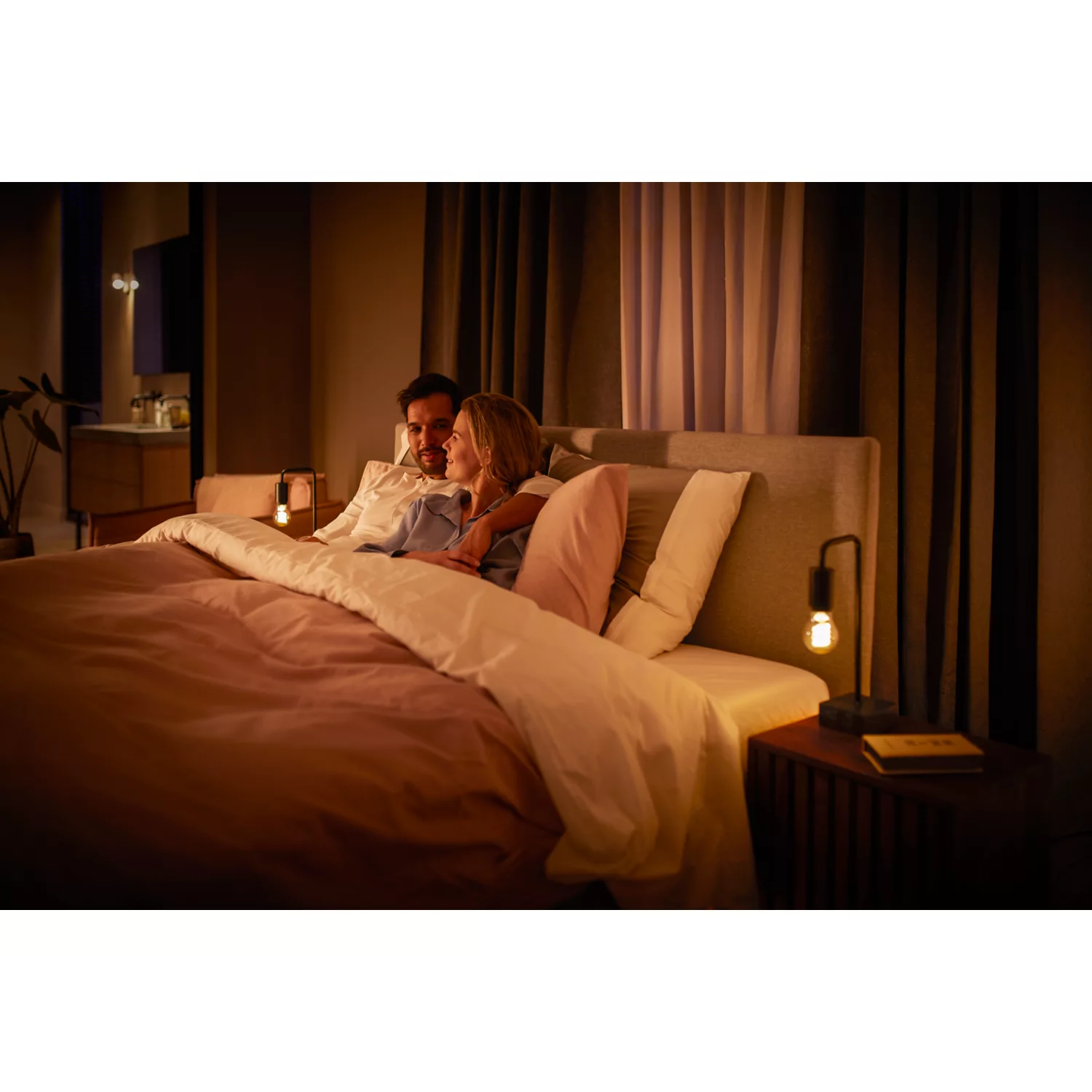 Couple in bed, under pink blanket, with side table lamp.