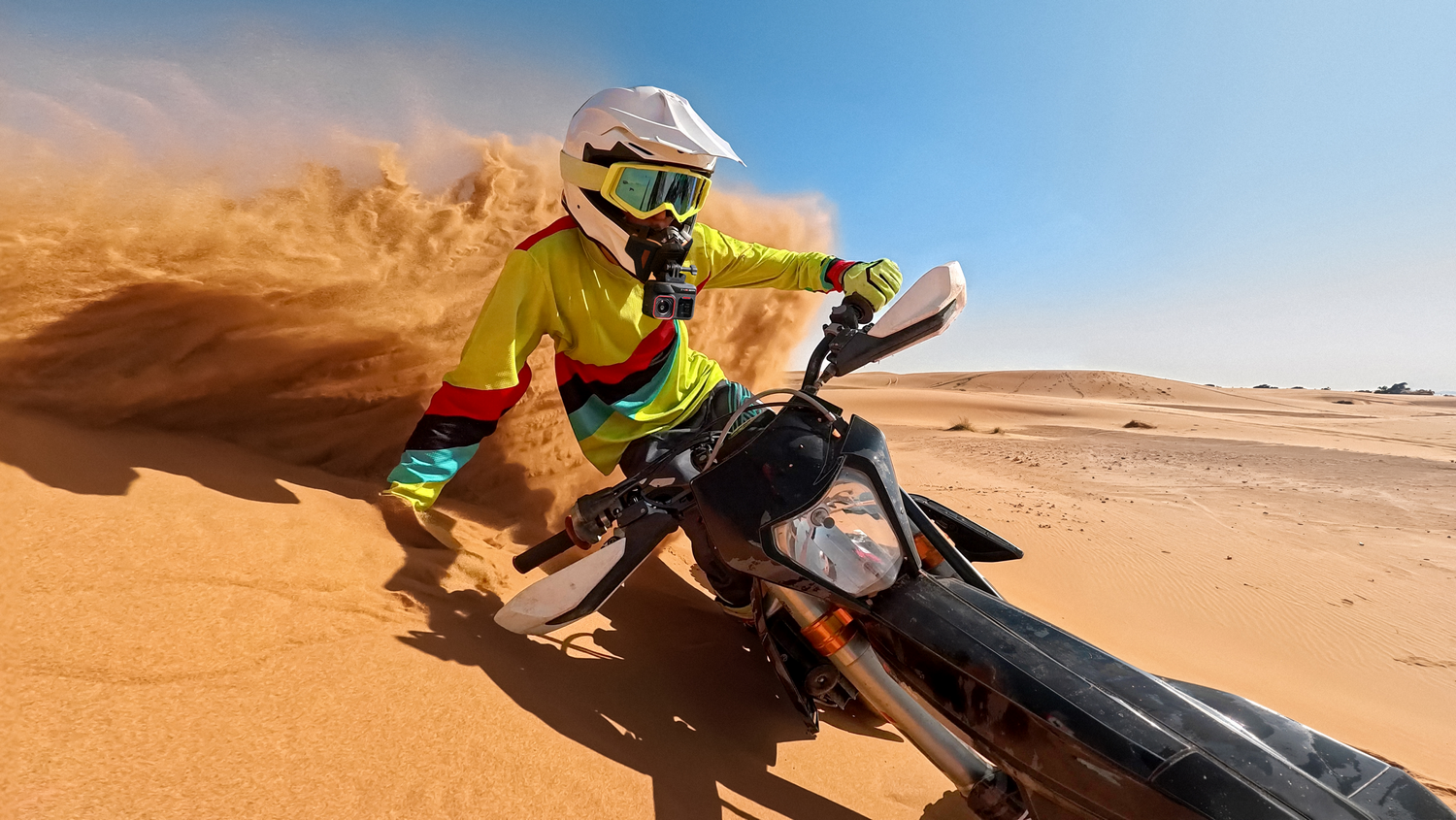 Una persona en una moto de cross, montando por la arena, con un fondo de cielo azul.