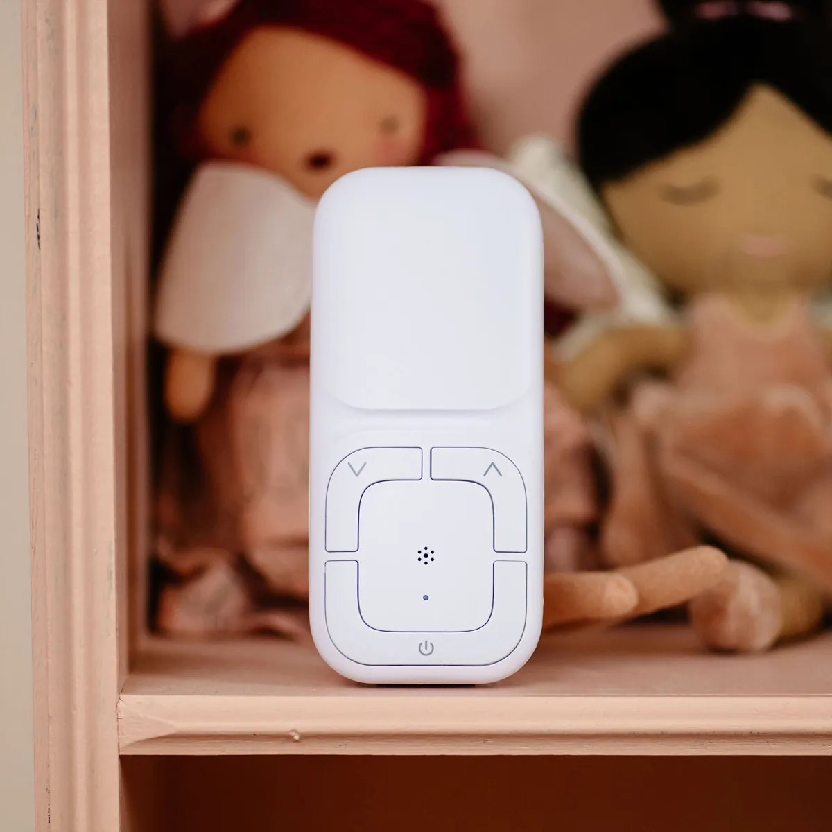 Un baby monitor bianco con pulsanti e un altoparlante è al centro. Bambole sullo sfondo, su uno scaffale rosa.