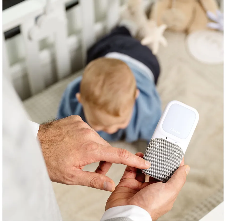 Un bébé dans un berceau. Une main tient un babyphone, pointant dessus. Lit et jouets en arrière-plan.