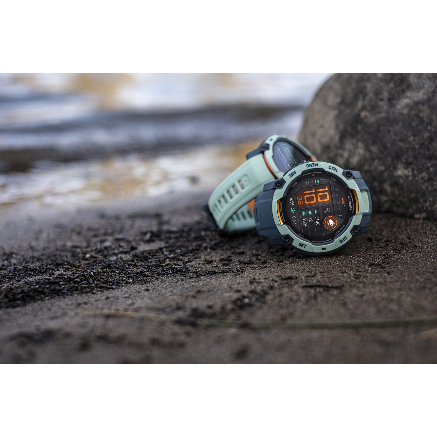 Twee smartwatches op een zandstrand bij water.