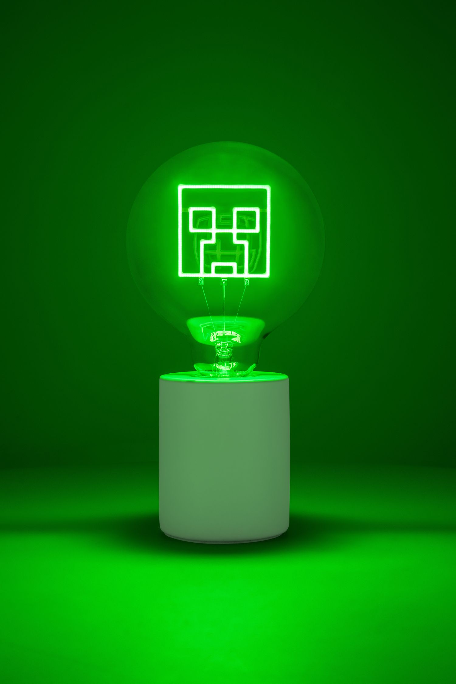 Egy zöld villanykörte egy Minecraft Creeper arccal, fehér állványon, zöld háttérrel.