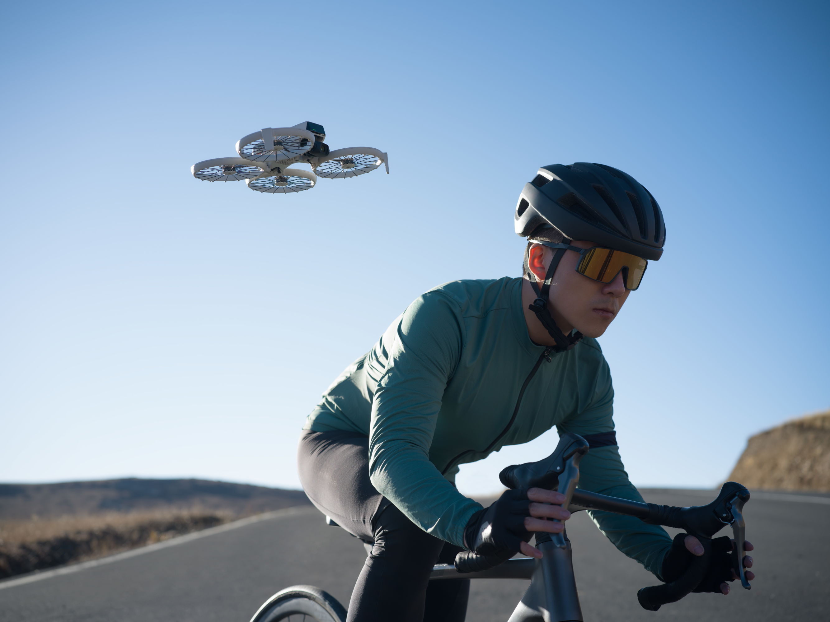 Ciclista su strada con drone sopra, occhiali da sole, cielo blu.