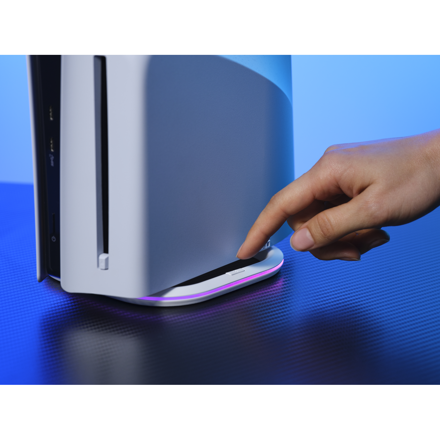 A hand touches a white game console on a blue surface. The console has a purple light.