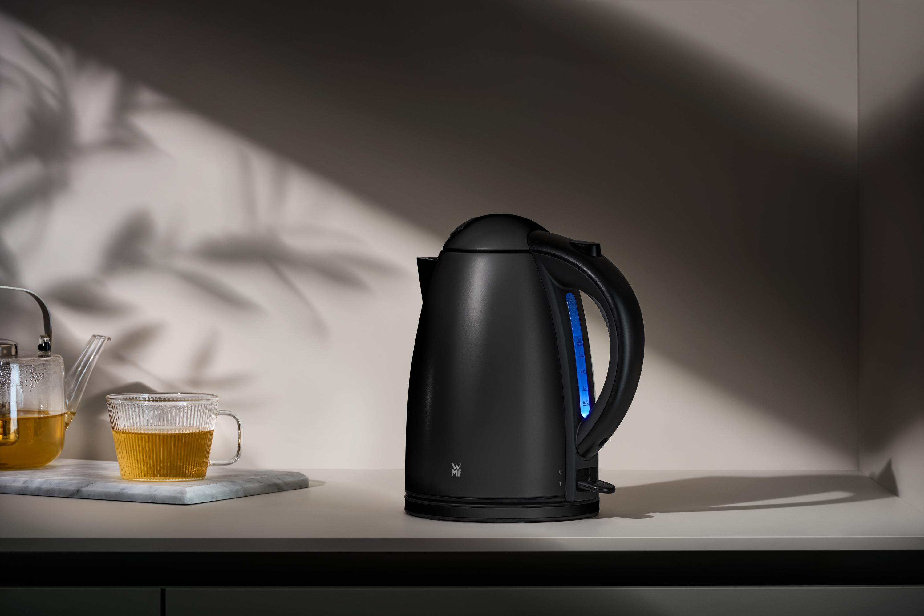 Ein schwarzer Wasserkocher mit Griff und blauem Licht. Er steht auf einer weißen Theke. Links ist eine Teetasse.