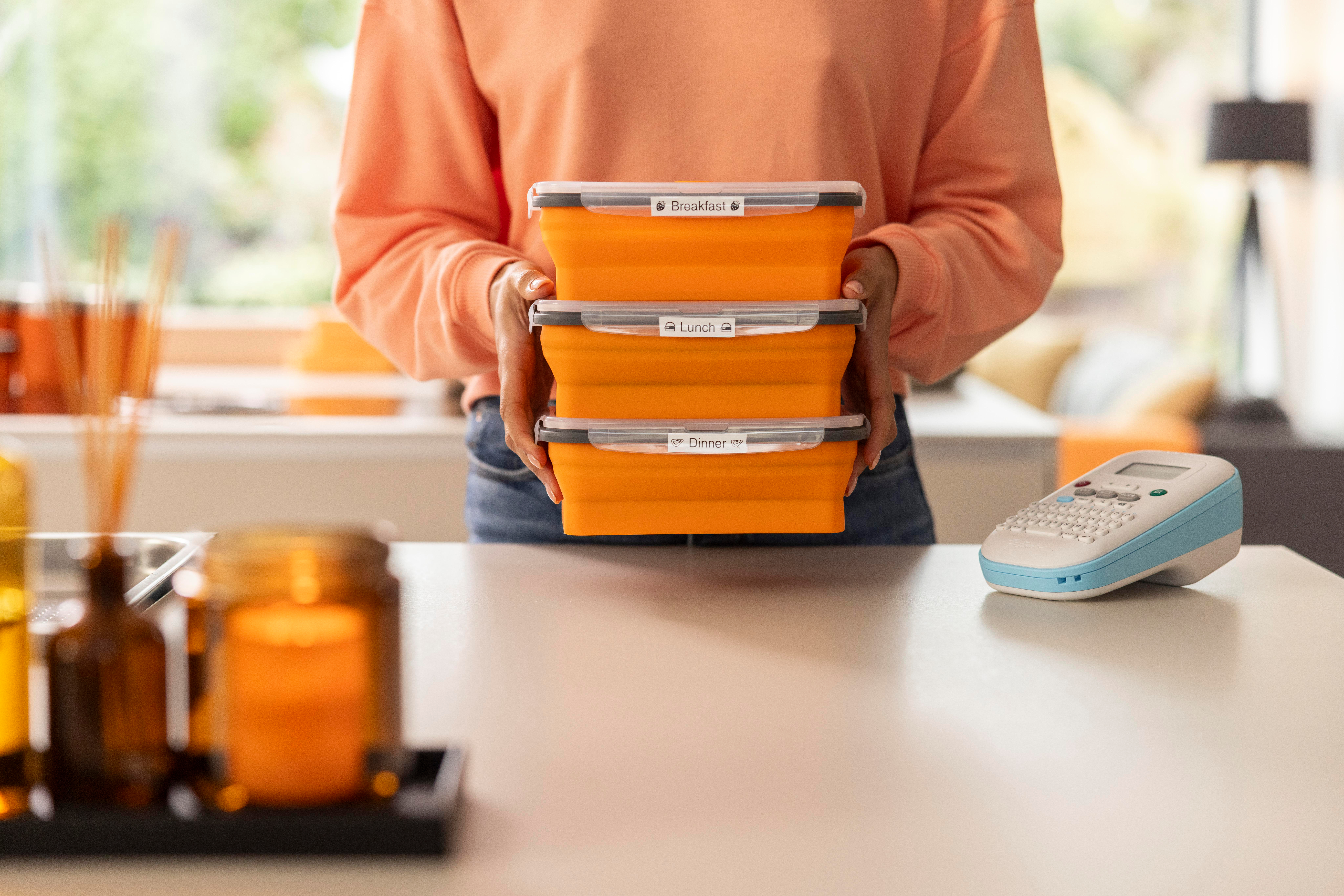 Eine Person, die drei orangefarbene Lunchboxen mit den Aufschriften Frühstück, Mittagessen und Abendessen hält.