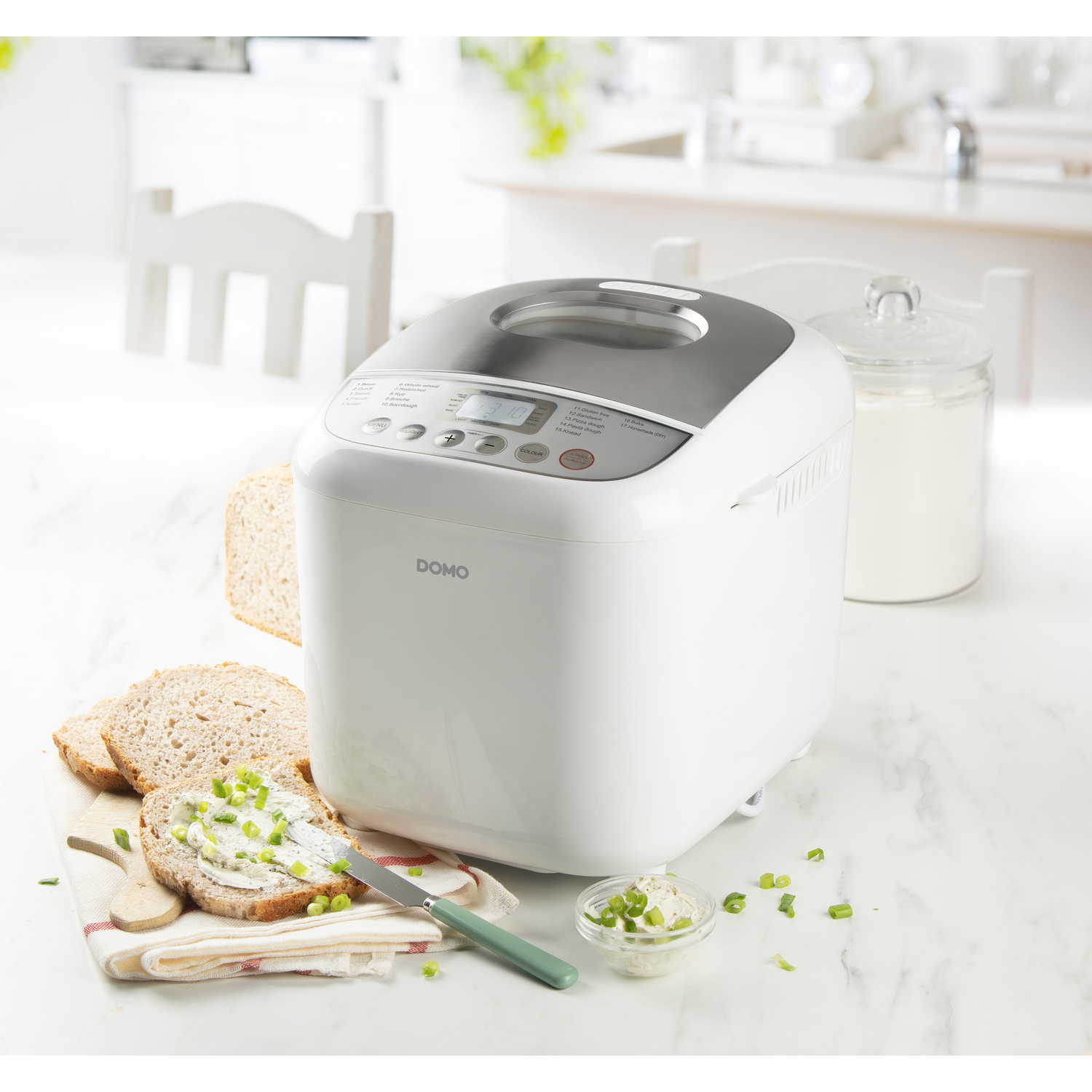 White bread maker next to bread slices with butter. A jar of flour and a knife are also present.