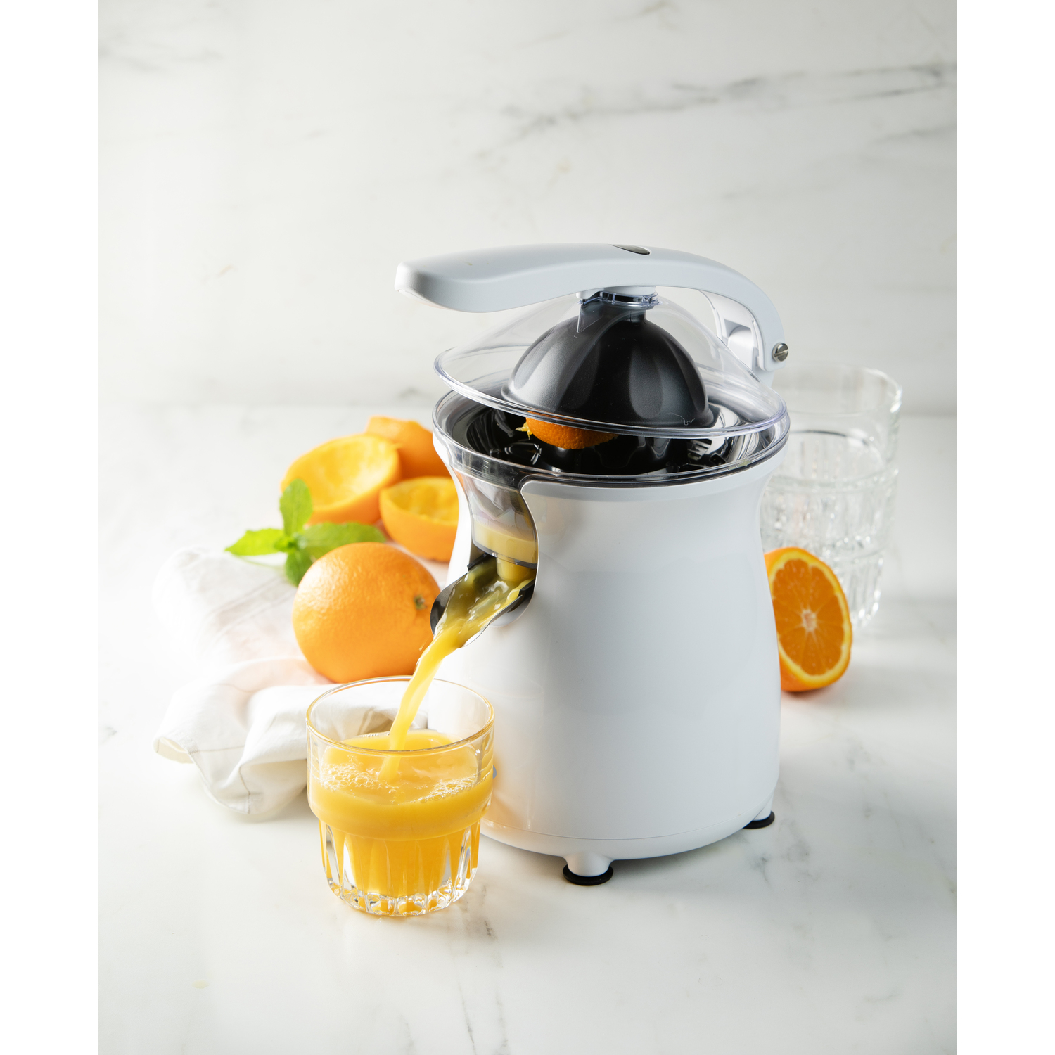 A white juicer with orange juice pouring into a glass. Oranges are on the table.