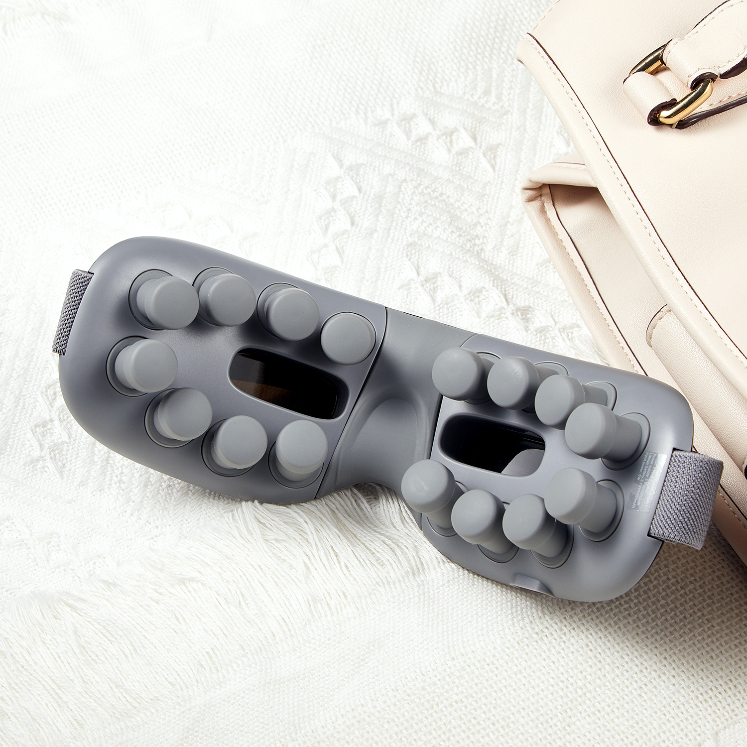 Gray eye massager on a white blanket near a purse. Shows the massager and bag.