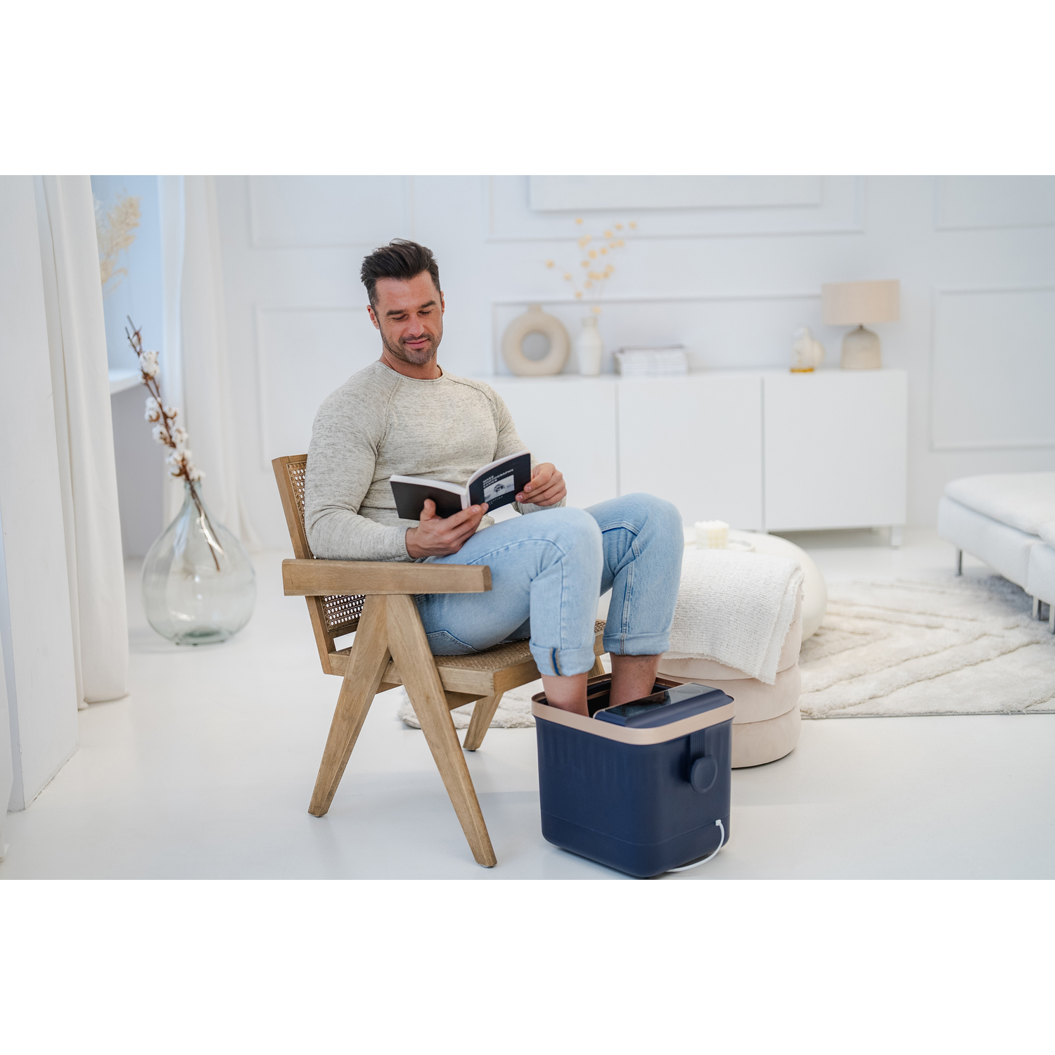 Man ontspant met een voetenbad. Leest een boek. Witte kamer, lichte achtergrond.