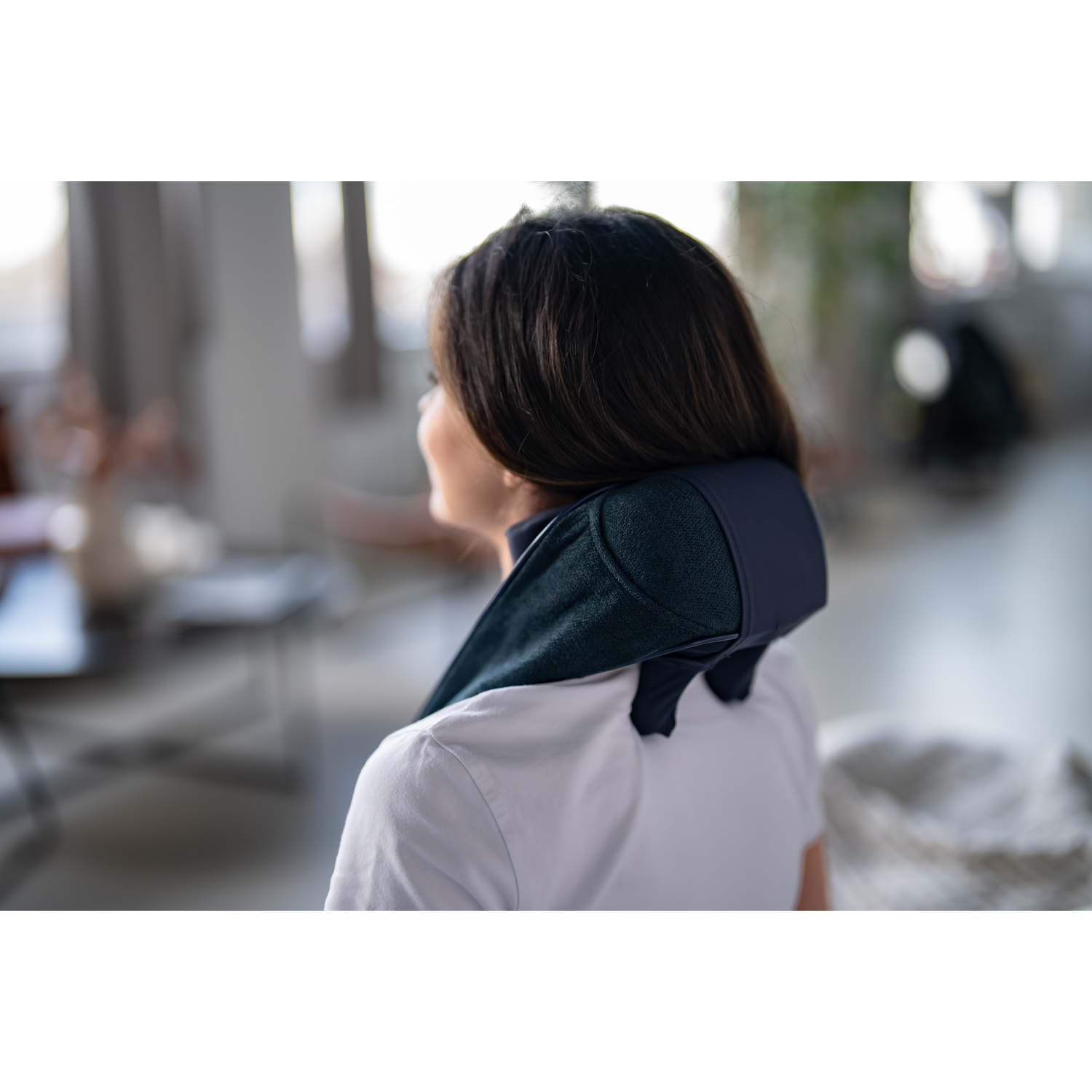 A woman's back with a neck massager. The massager is on her neck. The background is a room.