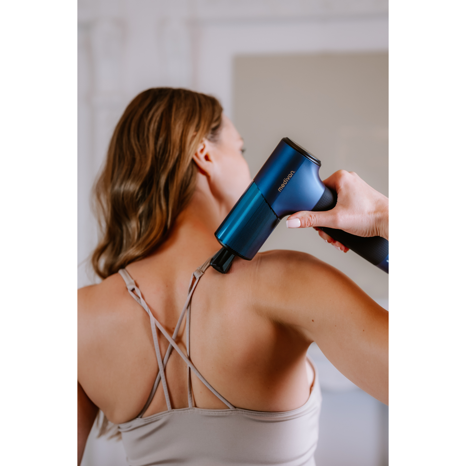 Woman is using massage gun on her back. Dark blue and black gun.