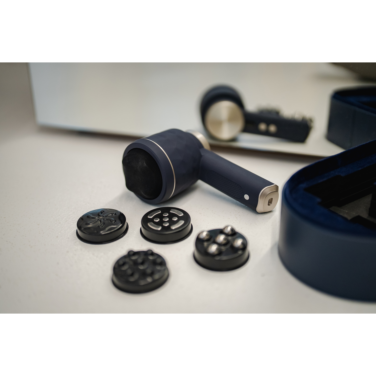 A navy blue massage device and its attachments are on a white surface. A mirror reflects the device. Accessories in a blue box.